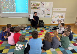 Dzieci siedzą w półkolu na dywanie, a prowadzący pokazuje im kartkę z symbolem CE i tłumaczy, co on oznacza podczas zajęć ABC Małego konsumenta.