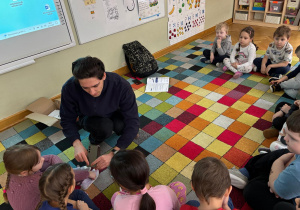 Dzieci siedzą w półkolu na dywanie, a prowadzący zajęcia pokazuje im znaczek „CE” na urządzeniu elektrycznym podczas zajęć ABC Małego konsumenta.