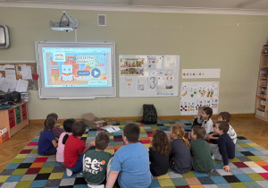 Dzieci siedzą w półkolu na dywanie i czekają aż rozpocznie się gra na tablicy interaktywnej podczas zajęć ABC Małego konsumenta.