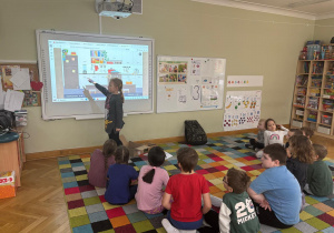 Dzieci siedzą na dywanie w półkolu, a dziewczynka dotyka ekranu tablicy interaktywnej i wskazuje przedmiot, który ma znaczek „CE” podczas zajęć ABC Małego Konsumenta.