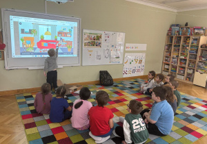 Dzieci siedzą na dywanie w półkolu, a chłopiec dotyka ekranu tablicy interaktywnej i wskazuje przedmiot, który ma znaczek „CE” podczas zajęć ABC Małego Konsumenta.