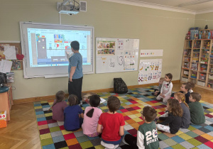 Dzieci siedzą na dywanie w półkolu, a chłopiec dotyka ekranu tablicy interaktywnej i wskazuje przedmiot, który ma znaczek „CE” podczas zajęć ABC Małego Konsumenta.