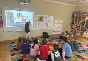 Dzieci siedzą na dywanie w półkolu, a dziewczynka dotyka ekranu tablicy interaktywnej i wskazuje przedmiot, który ma znaczek „CE” podczas zajęć ABC Małego Konsumenta.