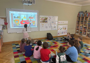 Dzieci siedzą na dywanie w półkolu, a dziewczynka dotyka ekranu tablicy interaktywnej i wskazuje przedmiot, który ma znaczek „CE” podczas zajęć ABC Małego Konsumenta.