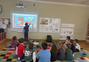 Dzieci siedzą na dywanie w półkolu, a dziewczynka dotyka ekranu tablicy interaktywnej i wskazuje przedmiot, który ma znaczek „CE” podczas zajęć ABC Małego Konsumenta.