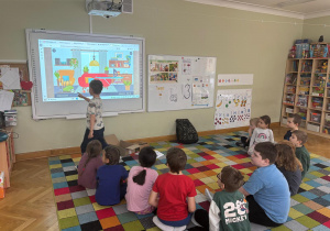 Dzieci siedzą na dywanie w półkolu, a chłopiec dotyka ekranu tablicy interaktywnej i wskazuje przedmiot, który ma znaczek „CE” podczas zajęć ABC Małego Konsumenta.