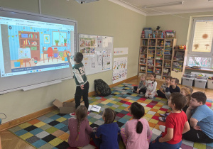 Dzieci siedzą na dywanie w półkolu, a chłopiec dotyka ekranu tablicy interaktywnej i wskazuje przedmiot, który ma znaczek „CE” podczas zajęć ABC Małego Konsumenta.