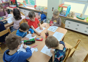 Dzieci siedzą przy stolikach i kolorują kredkami kolorowanki przyniesione przez jedną z mam.