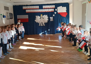 Dzieci ustawione w dwóch rzędach na przeciw siebie stoją na baczność i śpiewają hymn polski "Mazurek Dąbrowskiego", młodsze dzieci machają flagami.