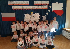 Starsza grupa pozuje do grupowego zdjęcia w galowych strojach na tle dekoracji z okazji 11 listopada.