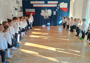 Dzieci ustawione w dwóch rzędach na przeciw siebie stoją na baczność i śpiewają hymn polski "Mazurek Dąbrowskiego".