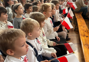 Dzieci ubrane na galowo siedzą na ławeczce z flagami biało-czerwonymi i oglądają występ innych uczestników wydarzenia.