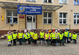 Dzieci ubrane w odblaskowe kamizelki stoją przed szkołą podstawową tuż po zakończeniu przeglądu pieśni patriotycznych.