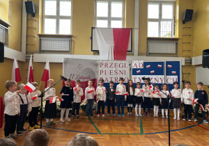 Dzieci ubrane na galowo z flagami biało-czerwonymi stoją ustawione w półkolu i wykonują przygotowaną na występ pieśń patriotyczną pt. "Szara Piechota".