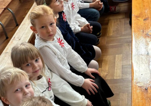 Dzieci ubrane na galowo siedzą na ławeczce i czekają na rozpoczęcie wydarzenia.