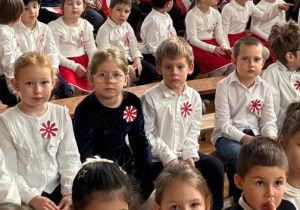 Dzieci ubrane na galowo siedzą na ławeczce i czekają na rozpoczęcie wydarzenia.