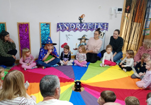 Nauczycielka w stroju czarodziejki siedzi wraz z dziećmi oraz ich rodzicami na chuście animacyjnej.