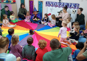 Nauczycielka w stroju czarodziejki siedzi wraz z dziećmi oraz ich rodzicami na chuście animacyjnej.