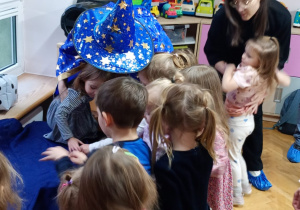 Dzieci stoją przy stoliku zasłoniętym obrusem i za pomocą dotyku rozpoznają przedmioty, które się pod nim kryją.