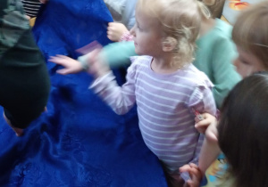 Dzieci stoją przy stoliku zasłoniętym obrusem i za pomocą dotyku rozpoznają przedmioty, które się pod nim kryją.