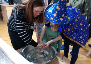 Chłopiec z pomocą nauczycielki trzyma klucz nad miską z wodą, a jego mama wlewa przez dziurkę wosk.