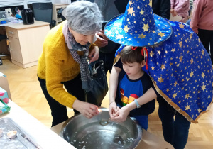 Chłopiec z pomocą nauczycielki trzyma klucz nad miską z wodą, a jego babcia wlewa przez dziurkę wosk.