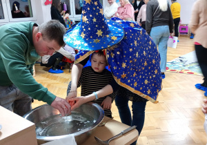 Chłopiec z pomocą nauczycielki trzyma klucz nad miską z wodą, a jego tata wlewa przez dziurkę wosk.