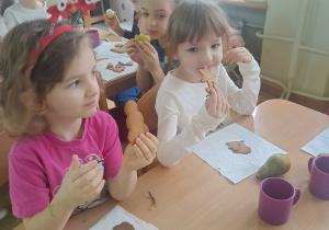 Dziewczynki siedzą przy stolikach i jedzą wykonane samodzielnie pierniczki z gruszką na podwieczorek.