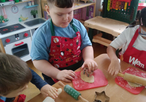 Chłopcy i dziewczynka siedzą przy stoliku, rozwałkowują ciasto na pierniczki i wyciskają wybrane przez siebie wzory przy użyciu foremek.
