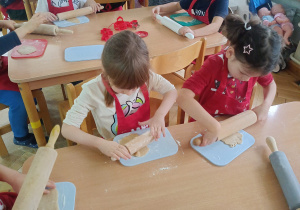 Dzieci siedzą przy stoliku, rozwałkowują ciasto na pierniczki i wyciskają wybrane przez siebie wzory przy użyciu foremek.