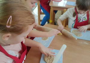 Dzieci siedzą przy stoliku, rozwałkowują ciasto na pierniczki i wyciskają wybrane przez siebie wzory przy użyciu foremek.