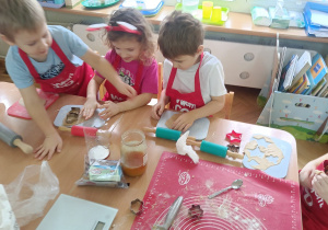 Dzieci siedzą przy stoliku, rozwałkowują ciasto na pierniczki i wyciskają wybrane przez siebie wzory przy użyciu foremek.