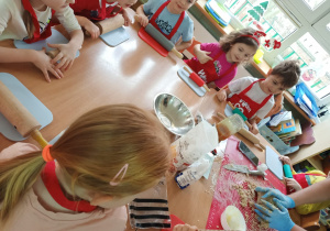 Dzieci siedzą przy stolikach i przygotowują się do robienia pierniczków na zajęciach kulinarnych.