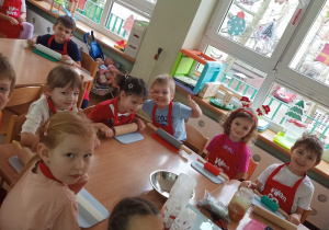 Dzieci siedzą przy stolikach i przygotowują się do robienia pierniczków na zajęciach kulinarnych.