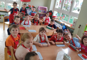 Dzieci siedzą przy stolikach i przygotowują się do robienia pierniczków na zajęciach kulinarnych.