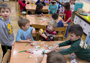 Dzieci siedzą przy stolikach i dekorują drewniane i styropianowe dekoracje świąteczne wg. własnego pomysłu.