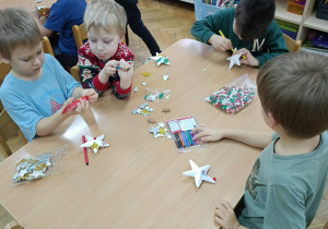 Dzieci siedzą przy stolikach i markerami dekorują drewniane i styropianowe dekoracje świąteczne wg. własnego pomysłu.