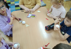 Dzieci siedzą przy stolikach i markerami dekorują drewniane i styropianowe dekoracje świąteczne wg. własnego pomysłu.
