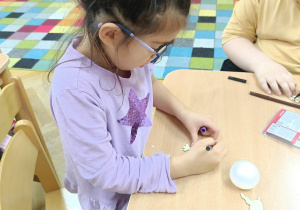 Dziewczynka siedzi przy stoliku i markerami dekoruje drewnianą dekoracje świąteczną wg. własnego pomysłu.