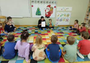 Dzieci siedzą na dywanie w półkolu i oglądają książeczkę prezentowaną przez mamę dziewczynki, a przygotowaną przez ich koleżankę.