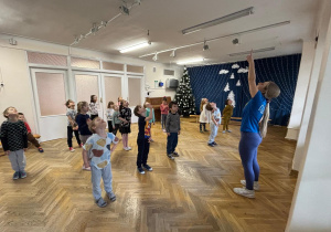 Dzieci stoją w rozsypce na sali gimnastycznej i wykonują rozgrzewkę na zajęciach z zumby podnosząc głowy do góry.