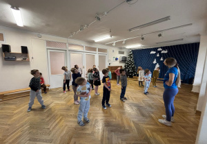 Dzieci stoją w rozsypce na sali gimnastycznej i wykonują rozgrzewkę na zajęciach z zumby pochylając głowy do boku.