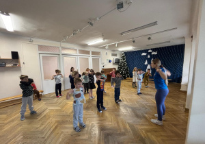 Dzieci stoją w rozsypce na sali gimnastycznej i wykonują krążenia ramionami na zajęciach z zumby.