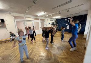 Dzieci stoją w rozsypce na sali gimnastycznej i wykonują podskoki z rękami uniesionymi do góry na zajęciach z zumby.