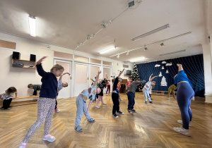 Dzieci ustawione w rozsypce wykonują skłony w bok wspólnie z prowadzącą zajęcia z zumby.