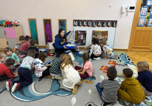 Dzieci siedzą na dywanie i podzielone na małe zespoły czekają aż prowadząca przydzieli każdej grupie pudełko z klockami.