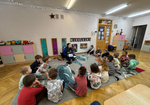 Dzieci siedzą na dywanie w półkoli i słuchają prowadzącej, czym będą się zajmować na zajęciach.