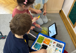 Dziewczynka i dwóch chłopców buduje konstrukcję z klocków wg instrukcji wyświetlanej na tablecie.