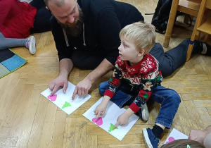 Chłopiec z tatą wskazuje poszczególne obrazki na kartce wg. instrukcji nauczycielki.