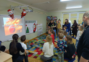 Rodzice z dziećmi stoją i wspólnie śpiewają piosenkę wyświetlaną na tablicy multimedialnej podczas zabawy karaoke.